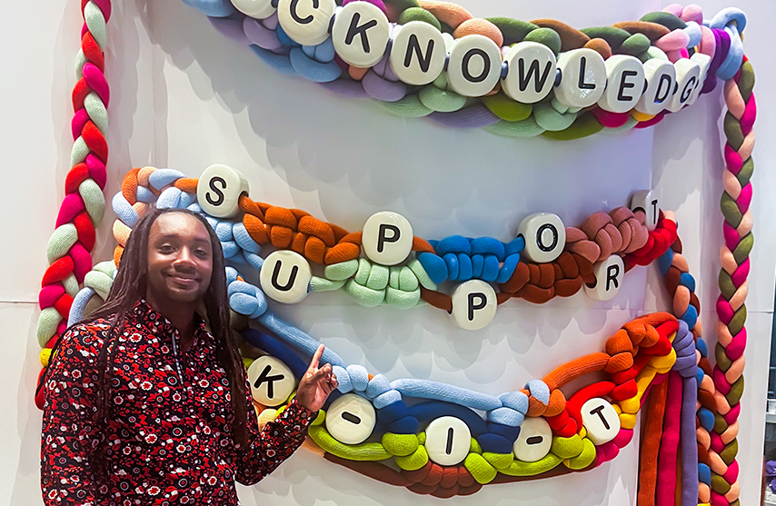 Tre Moss '25 MSW smiles while posing in front of large decorations meant to look like childhood beaded friendship bracelets.