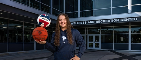 Carrie Hungate, '28, stands in front of the Wellness and Recreation Center at St. Ambrose University and smiles into the camera.