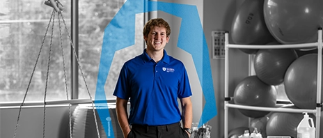 Ivan Hovland smiles into the camera while standing in an Occupational Therapy classroom. He smiles into the camera while a blue Ambrosian arc surrounds him.