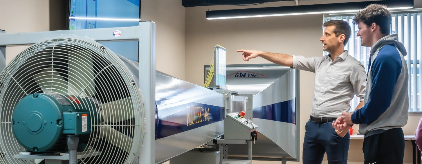 An engineering student and professor with a large wind tunnel in the background.