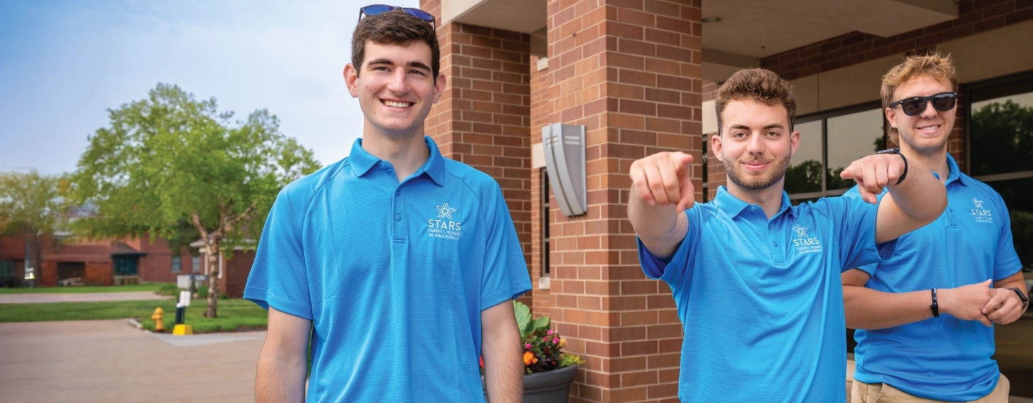 Three students in matching blue shirts, outdoors, with one in the middle pointing at the camera.